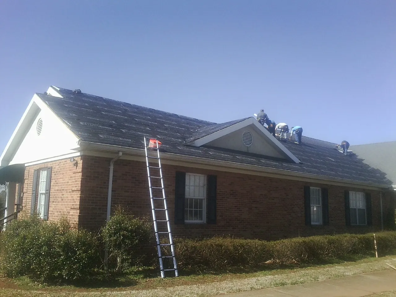 Crew tearing shingles off a brick ranch roof with a ladder set — Emergency Roof Repair work by our crew in Utica