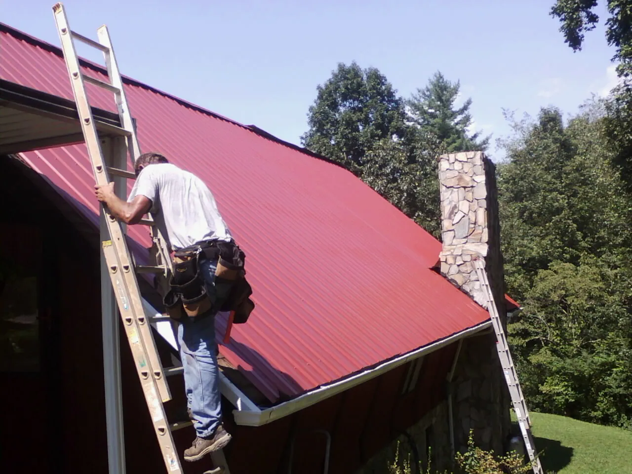 Roofer on a ladder beside a red metal roof and stone chimney — Soffit & Fascia Repair by Roofing Brothers in Utica